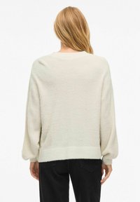 Kremo spalvos, per didelis megztinis su ilgomis rankovėmis, ribotais rankogaliais ir apačia. Minkšta, tekstūruota medžiaga su laisvu kirpimu. Be raštų ar akcentų.