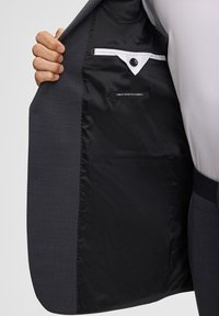 Ręka trzyma rozpiętą ciemną marynarkę, pokazującą czarne satynowe podszewki z zapiętą wewnętrzną kieszenią i metką z napisem "Najlepsza tkanina stretch".