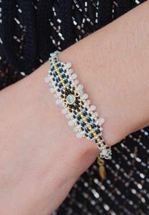 Bracelet en perles présentant des perles noires, dorées et transparentes avec un accent décoratif central, sur fond texturé.