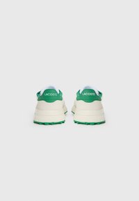 Lacoste sneakers met een crèmekleurige bovenkant, groene accenten en een witte kraag. Rond gevormd met een getextureerde buitenzool voor grip. Logo op de hak.