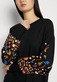 Sweatshirt noir à manches longues avec une broderie florale colorée dans des tons de bleu, orange et rose sur les manches. Col rond simple.