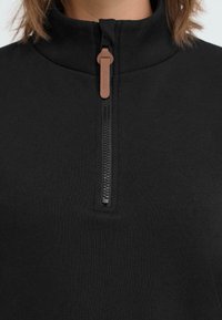 Zwarte pullover met een hoge kraag en een gedeeltelijke ritssluiting aan de voorkant. De rits heeft een bruine trekknop. Zachte, gladde stoftextuur.