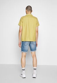 Camisa de botones amarilla de manga corta combinada con pantalones cortos de mezclilla. Los calcetines tienen rayas negras y unas zapatillas blancas completan el atuendo.