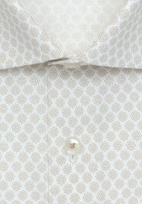 Chemise à col avec motif géométrique floral beige et boutons blancs sur un fond clair, tissu légèrement texturé et doux.