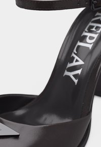 Μαύρο ανοιχτό πέδιλο με ψηλό τακούνι, λουράκι στον αστράγαλο και ασημένια τριγωνική λεπτομέρεια στο μπροστινό λουράκι.