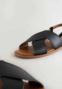 Sandali in pelle nera con design incrociato, punta aperta e suola in gomma marrone. Texture liscia con dettagli di cucitura a contrasto.