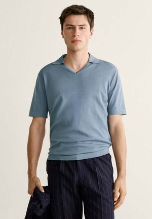 Un joven está de pie con una camisa polo de manga corta azul claro y pantalones oscuros con rayas finas, sosteniendo una chaqueta a juego en la mano izquierda.