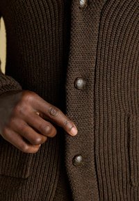 Barna kötött kardigán, texturált anyaggal, bordás mintázattal és díszgombokkal. Egy kéz kölcsönhatásba lép a gombos elülső résszel.