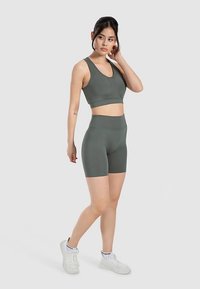 Olive green sports bra and matching high-waisted shorts, made from smooth, stretchy fabric. Model poses on a light gray background.