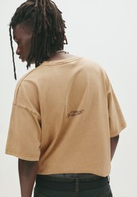 Brun oversized T-shirt i blødt bomuld med rund hals, der har et lille broderet logo på ryggen, korte ærmer og afslappet pasform.
