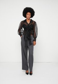 Blouse transparente à pois noirs avec des manches bouffantes, nouée à la taille, associée à un pantalon large gris et des talons aiguilles noirs.
