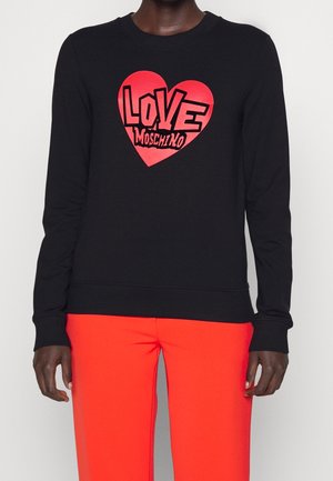 Personne portant un t-shirt noir à manches longues avec un cœur rouge et le texte "LOVE MOSCHINO", associé à un pantalon rouge vif.