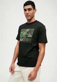 T-shirt en coton noir avec un graphique de crocodile vert et le texte "LACOSTE". Manches courtes, col rond, coupe décontractée. Texture lisse.