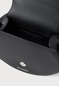 Μαύρη δερμάτινη τσάντα χειρός με λεία υφή, διαθέτοντας στρογγυλό μεταλλικό κούμπωμα και ανάγλυφη λεπτομέρεια λογότυπου στην μπροστινή πλευρά.