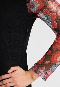 Vestido negro de encaje con un cuerpo texturizado y mangas transparentes con patrón floral en rojo, rosa y verde. Detalle de cremallera lateral visible.