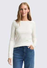Langsleeve geribbelde top in off-white met een wijde halslijn, gecombineerd met donkerblauwe jeans. De stof is soepel en lichtgewicht.