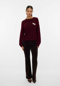 Maglione bordò in morbido knit con scollatura rotonda, caratterizzato da un dettaglio a forma di cuore di un leggero crema. Abbinato a pantaloni a vita alta marrone scuro.