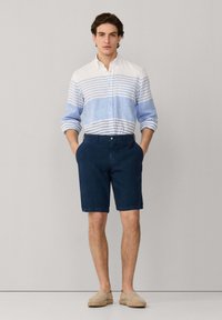 Hombre con camisa de manga larga a rayas blancas y azul claro, pantalones cortos azul marino y zapatos beige sin cordones, de pie con las manos en los bolsillos.