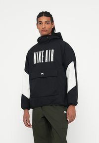 Juodas Nike Air pūkinis džemperis su dideliu baltu logotipu, baltais akcentiniais rankovėmis, priekinėmis kišenėmis ir reguliuojamu gobtuvu. Tekstūra lygi, medžiaga lengva.