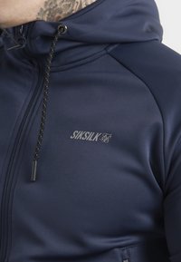 Marineblauwe, ritssluiting hoodie met een gladde textuur, verstelbare trekkoordcapuchon en een klein logo op de borst gedrukt.