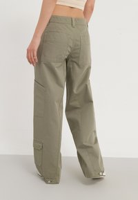 Pantalons cargo vert olive à jambes larges, avec de multiples poches et une texture en coton épais. Munis de fermetures à pression aux chevilles.