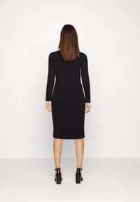 Femme aux cheveux bruns raides portant une robe noire ajustée, côtelée, à manches longues et des sandales à talons noires, debout face à un mur blanc.