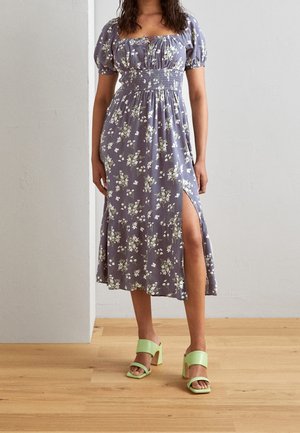 Femme portant une robe midi fleurie violette avec des manches bouffantes et une fente latérale, associée à des talons blocs verts clair ouverts à l'avant, sur un sol en bois.