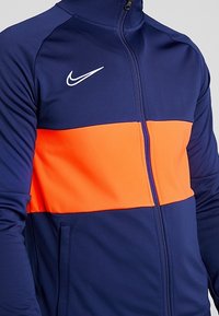 Veste de course Nike en bleu marine avec une bande horizontale orange sur la poitrine. Elle possède un col haut et une fermeture éclair à l'avant, fabriquée en tissu lisse et extensible.