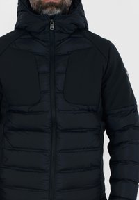 Giubbotto piumino nero con fronte trapuntato testurizzato, maniche in tessuto liscio, cappuccio e due ampi taschini sul petto; chiusura con zip.