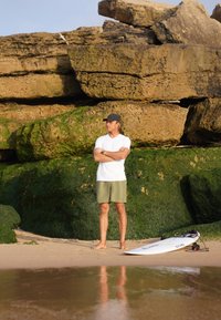 Uomo che indossa una camicia bianca a maniche corte e pantaloni corti verdi si trova su una spiaggia vicino a grandi rocce, con una tavola da surf appoggiata sulla sabbia.