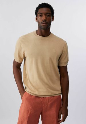 Uomo in piedi che indossa un maglione beige a maniche corte e pantaloni color ruggine con una mano in tasca, su sfondo semplice.