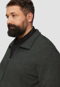 Dunkelgrüne elastische Jacke mit strukturiertem Strickmuster, ausgestattet mit einem großen Kragen und einem schwarzen Innenfutter.