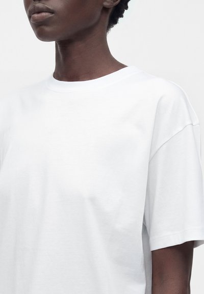 Adult with dark skin wearing a plain white short-sleeve crew neck T-shirt, shown from neck to mid-torso against a neutral background.
