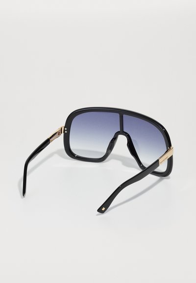 Black oversized sunglasses with gradient lenses, thick plastic frame, and gold-tone accents on the temples. Smooth texture throughout.