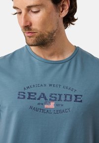 Jasnoniebieska bawełniana koszulka z nadrukiem "SEASIDE" w ciemnoniebieskim kolorze, z amerykańską flagą i współrzędnymi "45°N 123°W". Krótkie rękawy, okrągły dekolt.