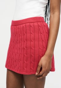 Γυναίκα που φοράει κόκκινη μίνι φούστα με πλεξούδες και λευκό αμάνικο τοπ, με το χέρι χαλαρό στο πλάι.