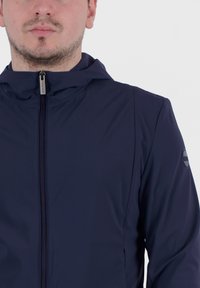 Giacca leggera di colore blu navy realizzata in tessuto morbido, con chiusura a zip, cappuccio e dettaglio del logo sulla manica sinistra.