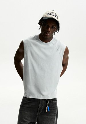 Un haut sans manches gris clair avec un col rond, porté avec un jean foncé. Une casquette crème avec le texte "VALLEY" est posée sur la tête. Des clés sont accrochées au jean.