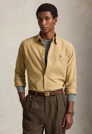 Jeune homme aux cheveux bouclés portant une chemise beige boutonnée sur un pull gris, un pantalon marron à carreaux et une ceinture en cuir marron avec une montre.