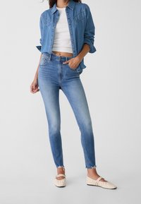 Kvinna i ljusblå högmidjade tight jeans, kort vit topp, denimskjorta och krämfärgade platta skor mot en enfärgad bakgrund.
