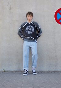 Tumehall pikkade varrukatega jakk valgete aktsentide ja logoga, helesinised ülekaalulised teksad ja mustad tossud valgete tallaga. Sile betoonist taust.