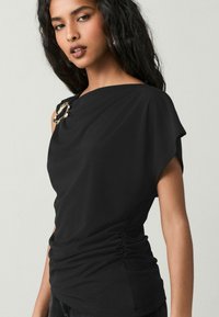 Top negru, cu umăr descoperit, cu un design drapat, având un detaliu de broșă metalică pe umărul stâng și textură pliată la talie.
