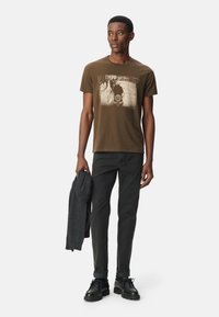 Camiseta de algodón marrón con un estampado gráfico, combinada con jeans oscuros y zapatos de cordones negros. El modelo sostiene un suéter gris.