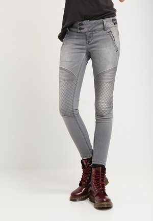 Jean skinny gris avec accents de texture matelassée aux genoux, présentant une fermeture à deux boutons et des détails cousus, associé à des bottes rouges à lacets.