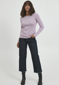 Pulover lung cu mânecă lungă de culoare mov deschis, din tricot cu nervuri, asortat cu blugi cropped de culoare închis. Modelul poartă cizme negre până la gleznă; părul este de lungime medie și drept.