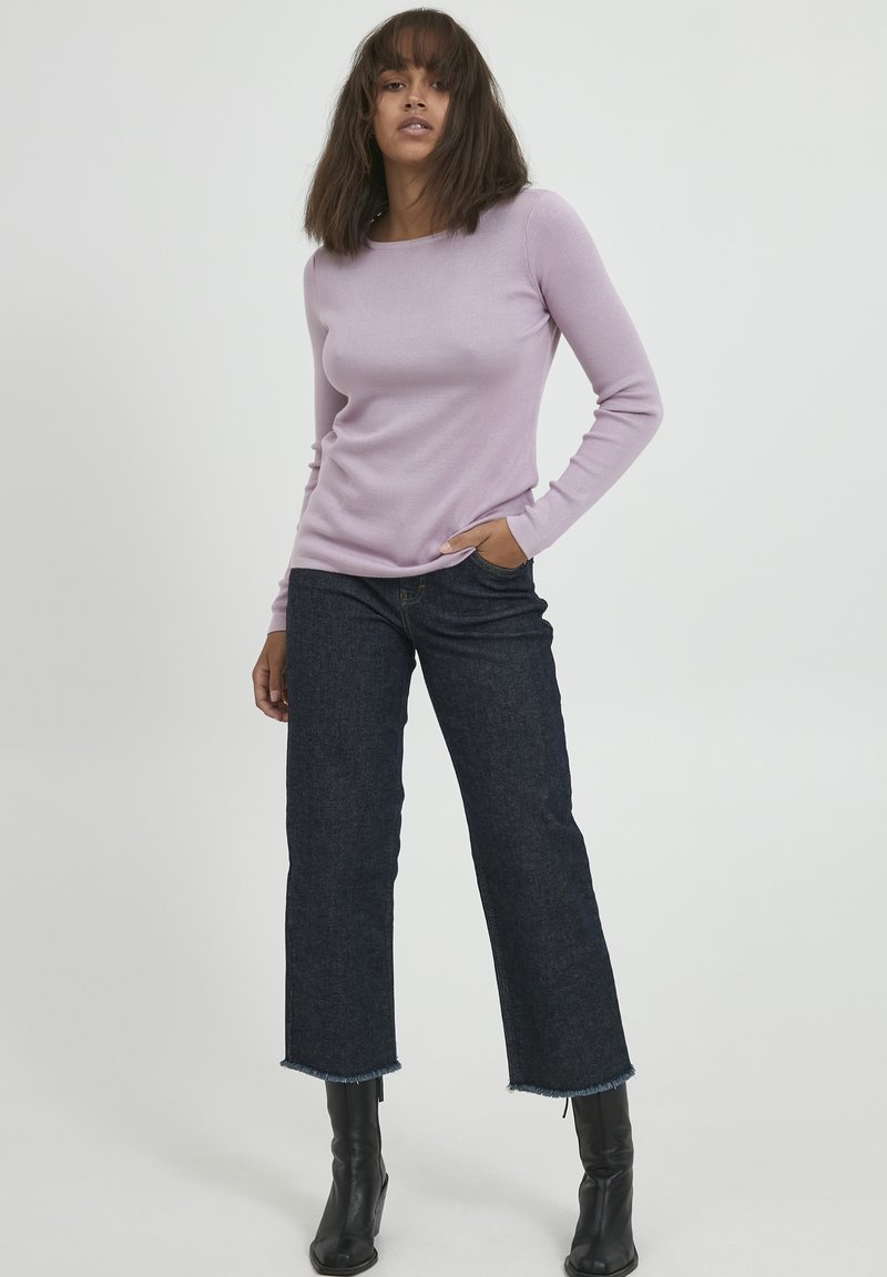 Pulover lung cu mânecă lungă de culoare mov deschis, din tricot cu nervuri, asortat cu blugi cropped de culoare închis. Modelul poartă cizme negre până la gleznă; părul este de lungime medie și drept.