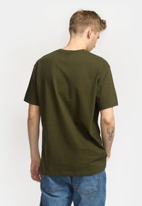Camiseta de algodón color caqui con cuello redondo y mangas cortas. La parte trasera presenta un diseño liso sin logos visibles.