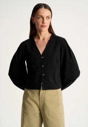 Kobieta ubrana w czarną bluzkę z długim rękawem i dekoltem w serek z trzema guzikami z przodu oraz beżowe spodnie z wysokim stanem, lekko patrzy na bok.