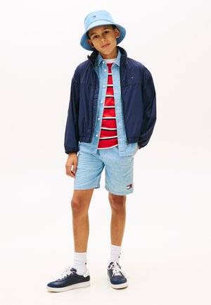 Jongen met een lichtblauwe vissershoed, marineblauw jasje, rood gestreept shirt, spijkershorts, witte sokken en marineblauwe sneakers, staand met één hand in zijn zak.