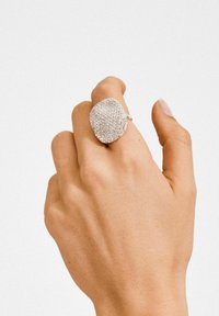 Hõberõngas tekstureeritud pinnaga, kaunistatud väikeste läbipaistvate kristallidega, ebaühtlaselt kujundatud, kantud helenaha tooniga käel.